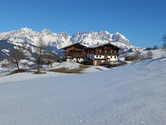 Hof Hinterkogl mit Hahnenkammblick Ein traditionelles Holzhaus in einer verschneiten Alpenlandschaft vor einem schroffen Gebirgsmassiv unter blauem Himmel.