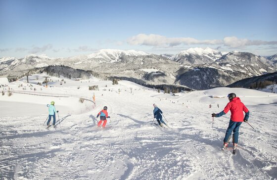 Skifahren auf der Steinplatte in Waidring Eine Gruppe von Skifahrern fährt den schneebedeckten Hang hinunter. Im Hintergrund sind majestätische Berge und ein klarer Himmel zu sehen.
