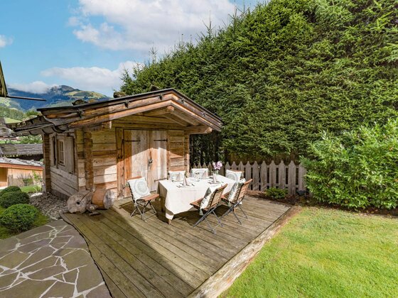 Gartenbereich Haus Schmidt Gedeckter Esstisch auf einer Holzterrasse vor einer kleinen Blockhütte im Garten.