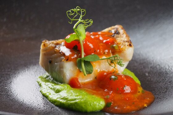Gebratenes Fischfilet auf Erbsenpüree mit Tomatensauce und Kräutergarnitur in einer dunklen Schale.