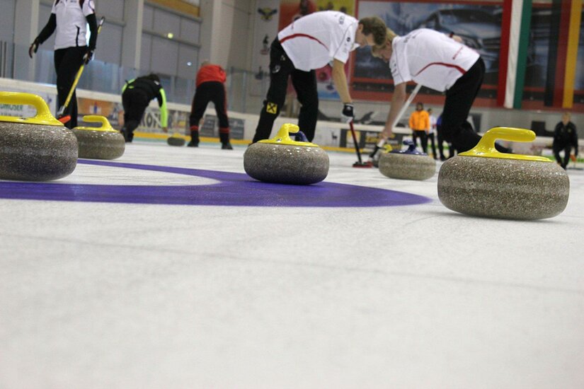 Mehrere Curlingsteine auf einer Eisfläche mit Spielern im Hintergrund.