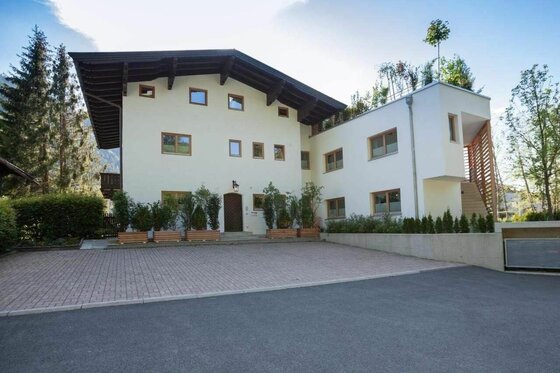 Hausansicht Kitz Apartment Vorderansicht eines mehrstöckigen Wohnhauses mit hellem Putz, dunklem Holzdach und gepflastertem Vorplatz.