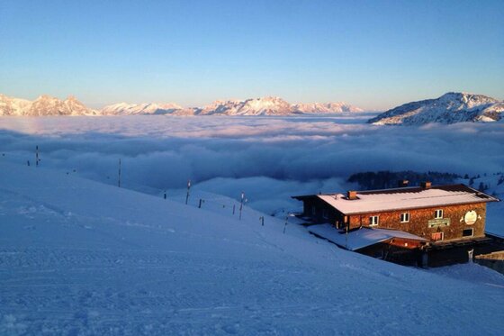 Hornköpflhütte im Winter Blick von einer verschneiten Skipiste auf die Hornköpflhütte über einer dichten Wolkendecke und Berggipfeln.