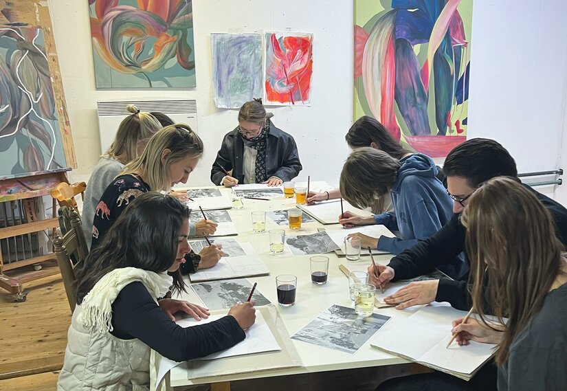 Céline Liebi Eine Gruppe von Künstlern sitzt an einem Tisch und zeichnet. Im Hintergrund sind bunte Gemälde zu sehen.