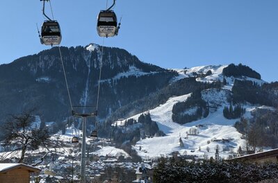 Gondelbahn vor Kitzbüheler Alpen Zwei Gondeln einer Seilbahn schweben vor einem verschneiten Bergmassiv mit Skipisten unter blauem Himmel.