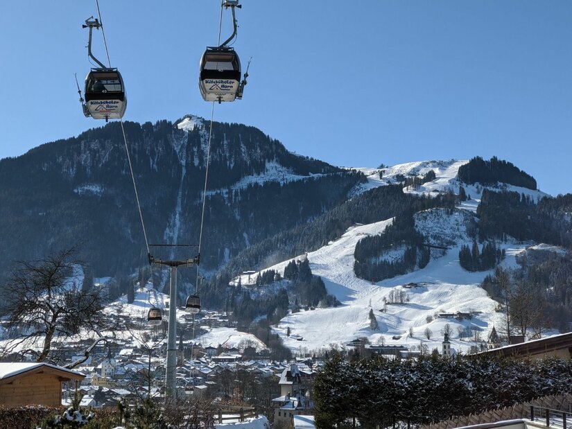 Gondelbahn vor Kitzbüheler Alpen Zwei Gondeln einer Seilbahn schweben vor einem verschneiten Bergmassiv mit Skipisten unter blauem Himmel.