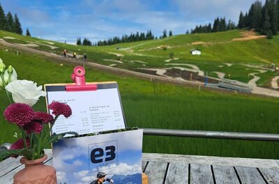 E3 Technik Lounge Holztisch mit Blumen und Speisekarte vor einem Bikepark im Hintergrund.