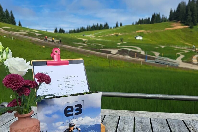 E3 Technik Lounge Holztisch mit Blumen und Speisekarte vor einem Bikepark im Hintergrund.