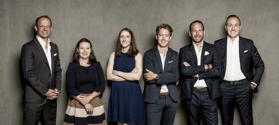 Team Cum Laude Immobilia Sechs Personen in Business-Kleidung stehen nebeneinander vor einer grauen Wand.