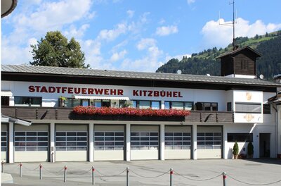 Ein Feuerwehrgebäude in Kitzbühel mit der Aufschrift "STADTFEUERWEHR KITZBÜHEL". Es gibt mehrere Fenster und einen klaren blauen Himmel im Hintergrund.