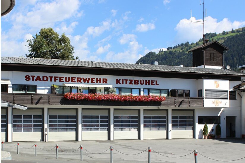 Feuerwehr Ein Feuerwehrgebäude in Kitzbühel mit der Aufschrift "STADTFEUERWEHR KITZBÜHEL". Es gibt mehrere Fenster und einen klaren blauen Himmel im Hintergrund.