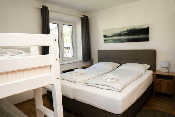 Ein gemütliches Schlafzimmer mit einem Doppelbett und einem Etagenbett. Große Fenster sorgen für viel Tageslicht.