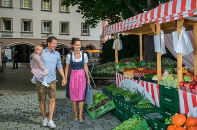 Kitzbüheler Genussmarkt Eine Familie besucht einen Wochenmarkt mit frischem Obst und Gemüse. Die Eltern halten ein Kind und genießen die gedeckte Marktbude.