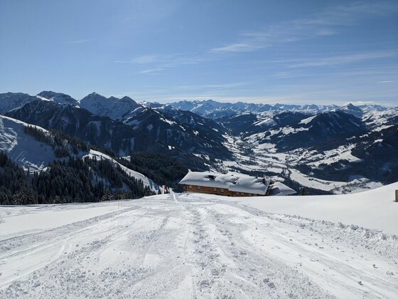 Bichlalm im Winter Verschneite Skipiste führt hinunter zu einer Holzhütte vor einem weiten Bergpanorama unter blauem Himmel.