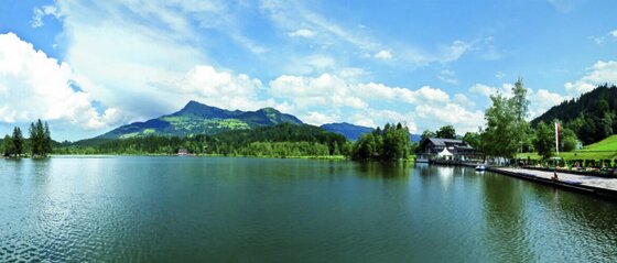 Panoramaaufnahme des Schwarzsees mit bewaldeten Bergen und einem Ufergebäude unter bewölktem Himmel.