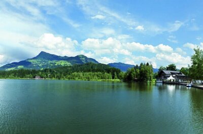 Panoramaaufnahme des Schwarzsees mit bewaldeten Bergen und einem Ufergebäude unter bewölktem Himmel.