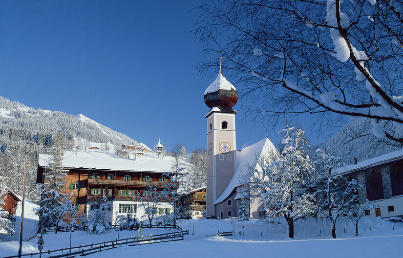 Verschneite Kirche mit Zwiebelturm und traditionelle Holzhäuser vor bewaldeten Bergen unter blauem Himmel.