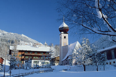 Verschneite Kirche mit Zwiebelturm und traditionelle Holzhäuser vor bewaldeten Bergen unter blauem Himmel.