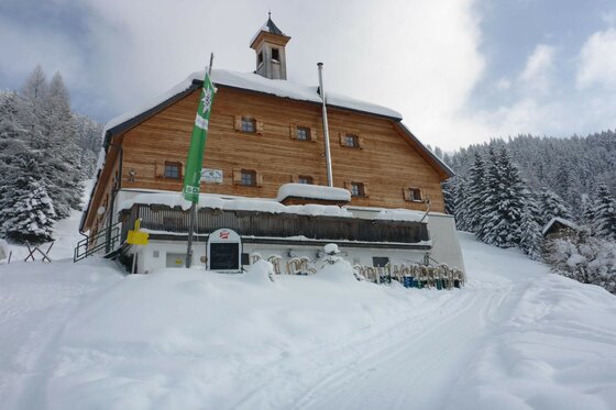 Verschneite Bochumer Hütte mit Holzfassade und kleiner Glocke auf dem Dach vor Nadelwald.