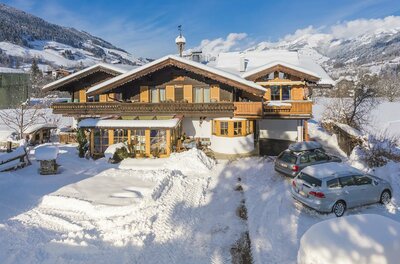 Landhaus Klotz Hausansicht Winter Alpines Holzhaus mit schneebedecktem Dach und zwei geparkten Autos im Vordergrund vor Bergkulisse.