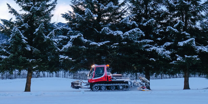 During the cross-country skiing event in Kitzbühel, a red Pistenbully grooms the trail through a snow-covered landscape dotted with fir trees.