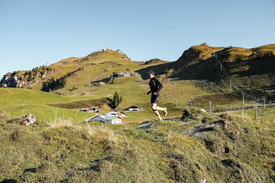Trailrunning in Kitzbühel Ein Trailrunner läuft über eine grasbewachsene Kuppe vor einer alpinen Bergkulisse mit Hütten und Liftanlagen.
