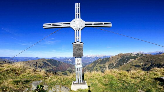 Gipfelkreuz am Gebra Trail Ein metallisches Gipfelkreuz auf einem grasbewachsenen Bergkamm vor blauem Himmel.