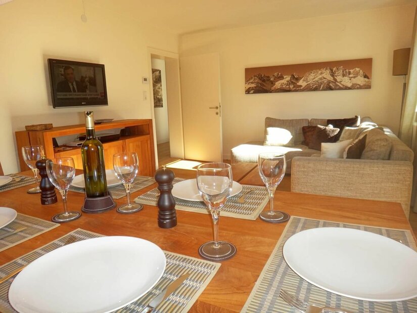Wohn-Esszimmer im Apartment Panorama Gedeckter Holztisch im Vordergrund, dahinter ein Sofa und ein Fernseher in einem hellen Wohnraum.