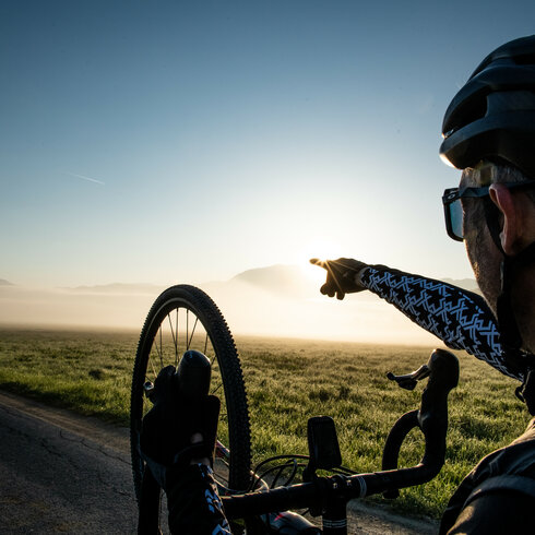 Ein Radfahrer zeigt auf die aufgehende Sonne über einer nebligen Landschaft, während er auf einer ruhigen Straße steht.