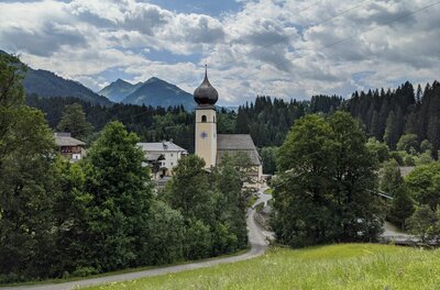 Kirche mit Zwiebelturm in Aurach vor bewaldeten Bergen und bewölktem Himmel.
