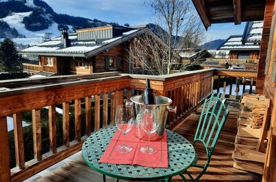 Balkon im Kitz Boutique Loft Holzbalkon mit rundem Tisch, zwei Weingläsern und Sektkühler vor alpiner Kulisse mit Chalets und Bergen.