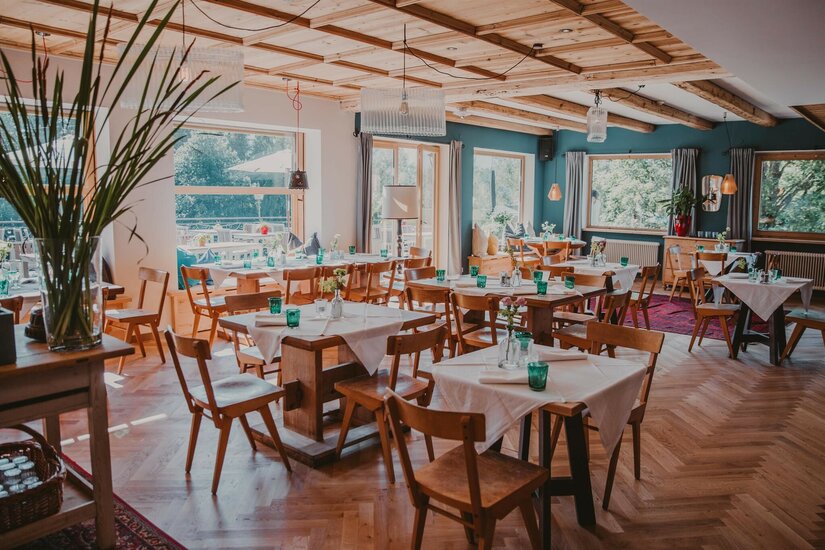 Innenansicht eines Restaurants mit Holztischen, Stühlen, Parkettboden und großen Fenstern.