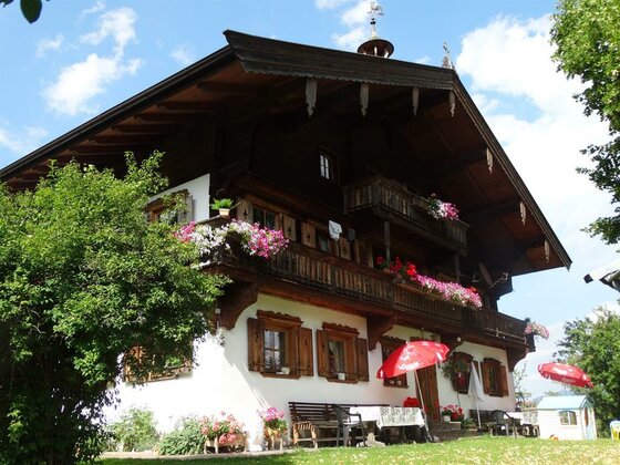 Alpenländisches Bauernhaus mit Holzbalkonen, Blumenschmuck und Außenterrasse unter blauem Himmel.