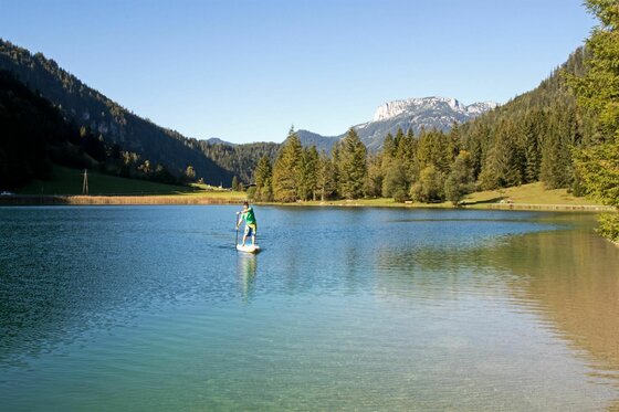 SUP Ein Mann paddelt auf einem Stand-Up-Paddleboard auf einem ruhigen Gewässer. Im Hintergrund sind grüne Berge und ein klarer blauer Himmel zu sehen.