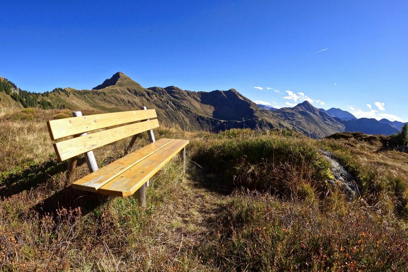 Rastplatz am Gebra Trail Holzbank auf einer Bergwiese vor einem Gebirgspanorama unter blauem Himmel.