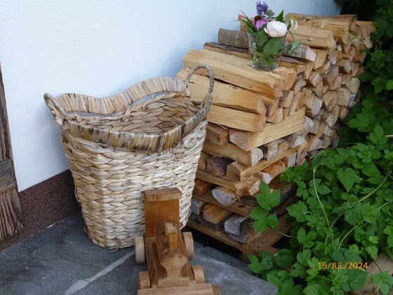 Garten 3 Ein geflochtener Korb, ein Holzspielzeug und aufgestapeltes Brennholz mit Blumenvase vor einer weißen Wand.