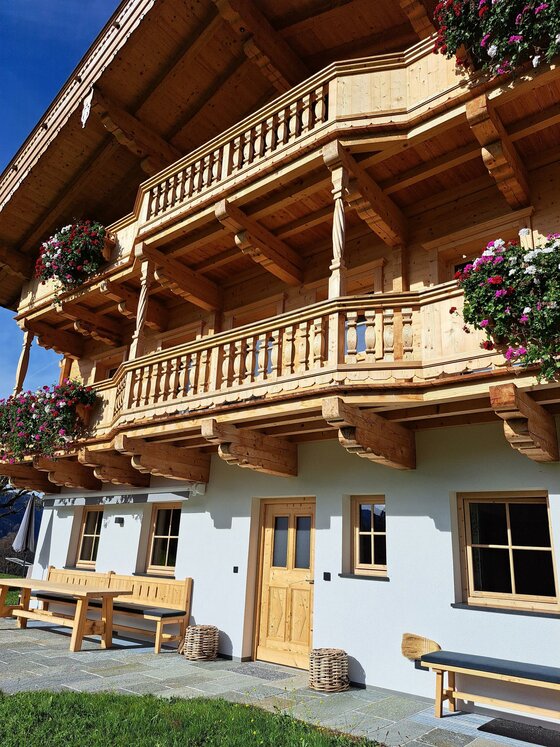 Ein rustikales Holzhaus mit einem Balkon, verziert mit blühenden Pflanzen. Die helle Fassade und die umgebende Natur schaffen eine einladende Atmosphäre.