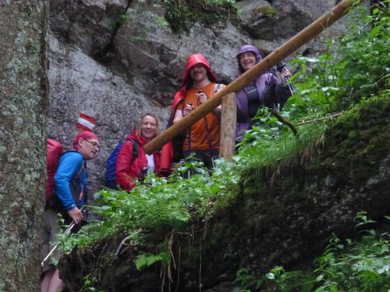 Wandergruppe bei Regen Vier Wanderer mit Regenbekleidung und Wanderstöcken auf einem schmalen Pfad an einer Felswand.