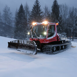 In Kitzbühel, a red Pistenbully is grooming the cross-country ski trail in a snow-covered landscape dotted with fir trees.