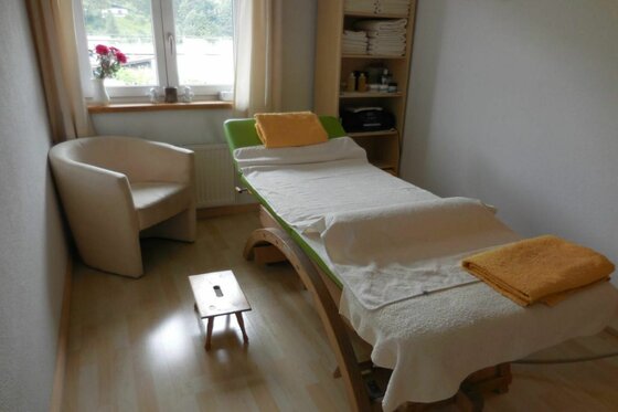 Behandlungsraum Wakolbinger Massageliege mit weißen und gelben Handtüchern in einem hellen Behandlungsraum mit Sessel und Fenster.