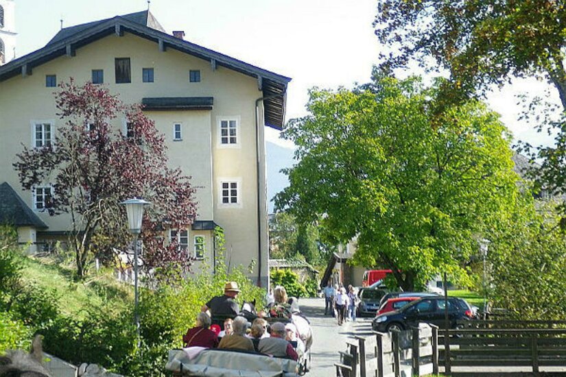 Pfarrhof Eine ruhige Straße mit einem großen Gebäude im Hintergrund. Menschen sitzen in einem offenen Fahrzeug und genießen die Landschaft.