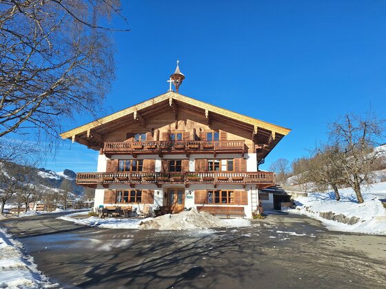 20260116_105453 Ein traditionelles Chalet in den Alpen mit einem Holzfassade. Es ist schneebedeckt und es scheint ein klarer blauer Himmel zu sein.