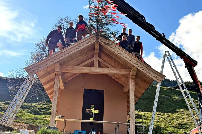 Personengruppe auf dem Dachstuhl einer neu gebauten Holzkapelle mit Richtbaum.