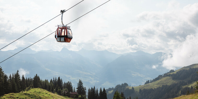 Eine rote Seilbahn der Hahnenkammbahn schwebt über die grünen Hänge des Hahnenkamms in Kitzbühel, umgeben von Bergen und einem bewölkten Himmel.