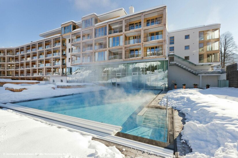 Wellnessbereich Kempinski Alpine Spa Dampfender Außenpool eines modernen Hotelgebäudes in einer verschneiten Winterlandschaft.