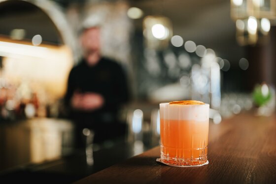 Ein orangefarbener Cocktail mit Schaumkrone auf einem Holztresen vor unscharfem Bar-Hintergrund.