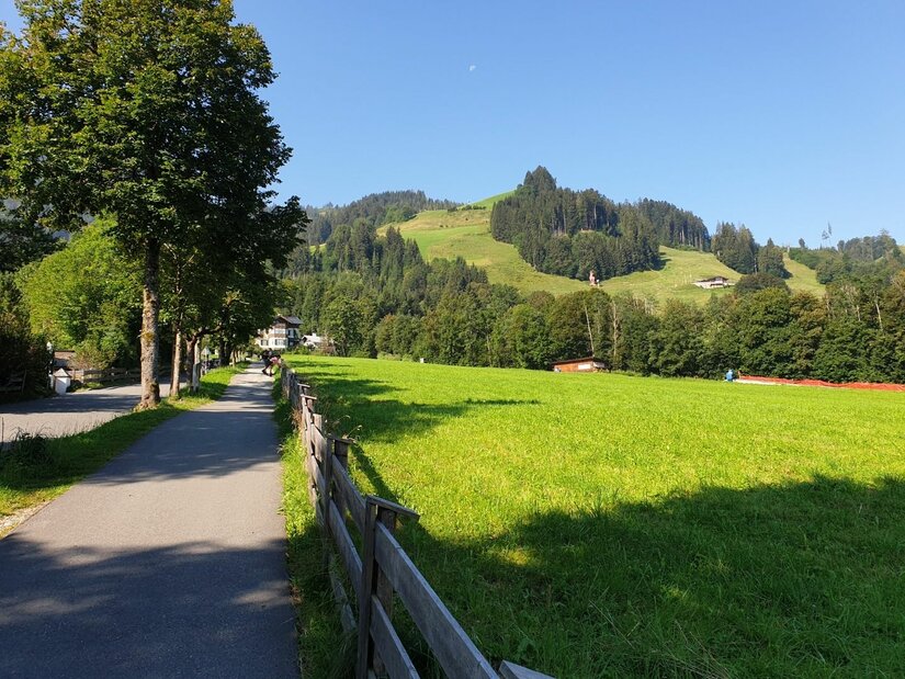Wanderweg am Kreuzweg Asphaltierter Wanderweg neben einer grünen Wiese mit Holzzaun und bewaldeten Hügeln im Hintergrund.