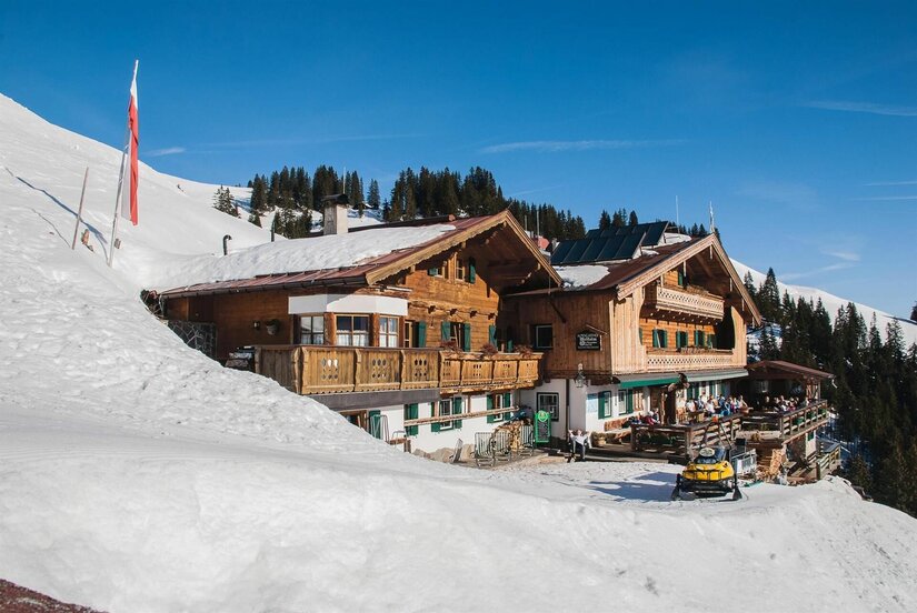 Melkalm Winteransicht Alpiner Berggasthof aus Holz in einer verschneiten Winterlandschaft unter blauem Himmel.