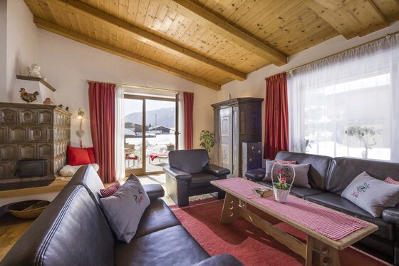 Wohnzimmer im Haus Skida Wohnraum mit Ledersofas, Holzbalkendecke, Kachelofen und Blick auf eine verschneite Terrasse.