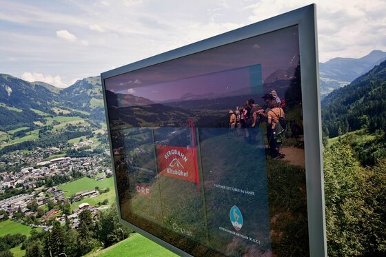 Infomonitor an der Streif Ein Monitor mit Informationen der Bergbahn Kitzbühel vor einer alpinen Bergkulisse mit Blick auf ein Tal.